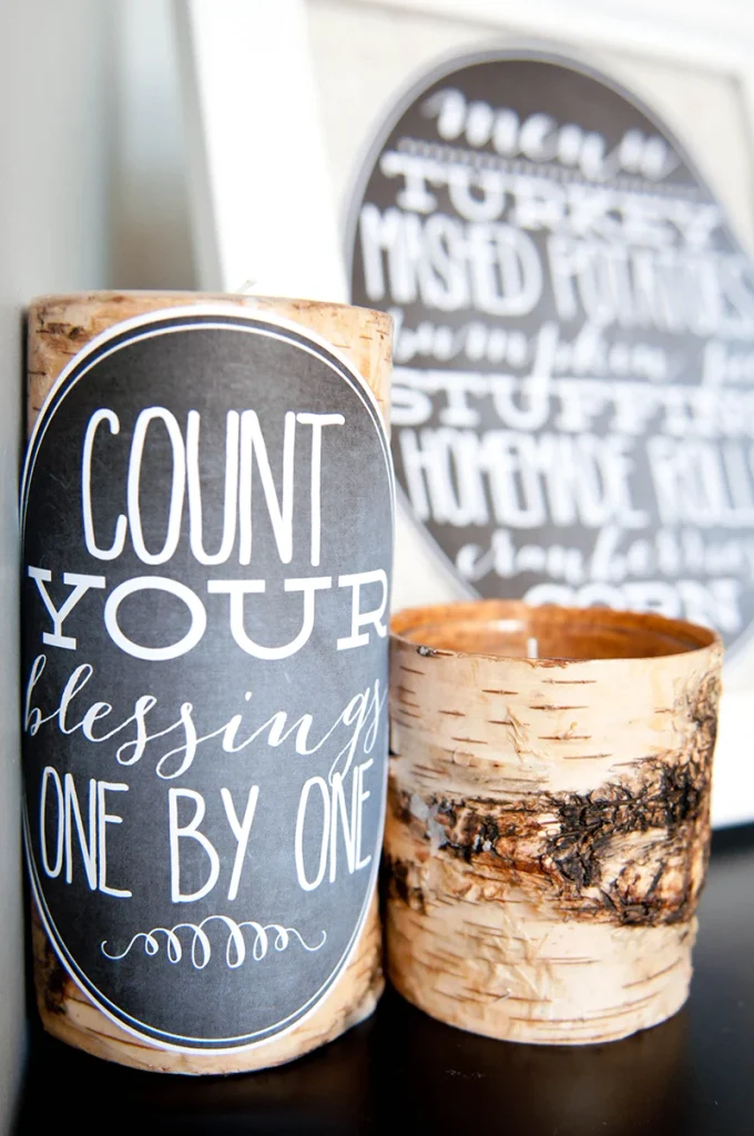 A thick birch bark pillar candle featuring a black oval label with the white text "COUNT YOUR blessings ONE BY ONE". A smaller birch candle sits next to it, and a blurred white frame with a menu can be seen in the background.
