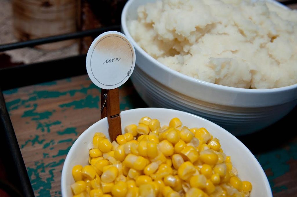 A close-up of a white bowl filled with corn on a Thanksgiving buffet table, featuring a printable round food label reading "corn" clipped to the side with a wooden clothespin next to a bowl of mashed potatoes.