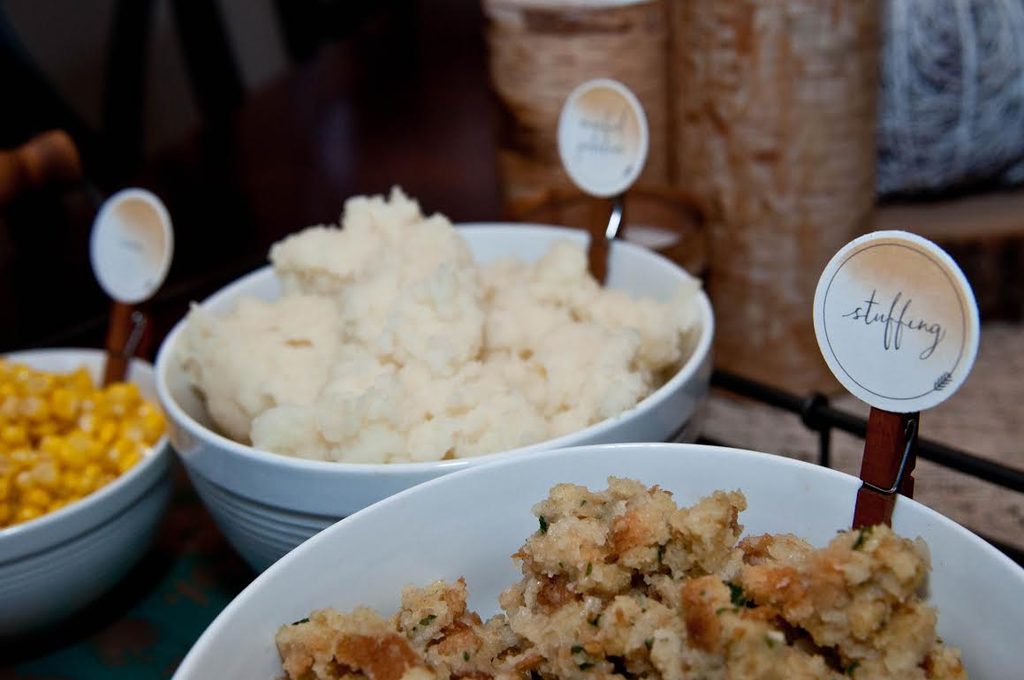 Several white serving bowls filled with Thanksgiving side dishes like stuffing, mashed potatoes, and corn on a buffet table, each labeled with round DIY food tags including "stuffing" held up by wooden clothespin clips.